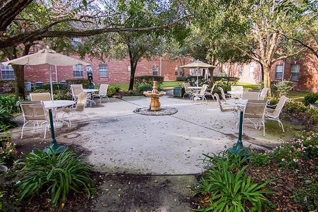 Outdoor courtyard area with several white metal tables and chairs, some shaded by umbrellas, surrounding a central fountain. The courtyard is bordered by green plants and trees, with a red brick building in the background.