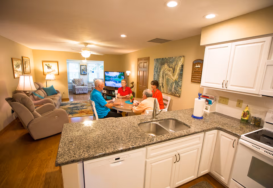 A cozy living area in a senior living facility with a kitchen counter in the foreground. Four elderly people are seated around a small round table, engaged in conversation. The room features a comfortable sofa, armchairs, a TV, and warm lighting with lamps and ceiling fan lights.