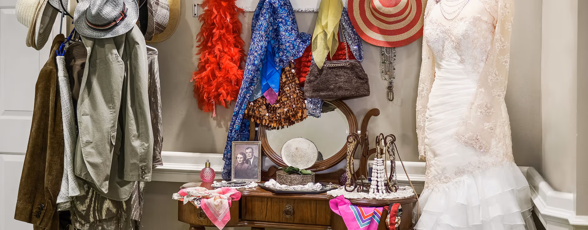 A vintage-style dressing area with a wooden vanity table holding various accessories including scarves, jewelry, a framed black-and-white photo, and a round mirror. Above the table, colorful scarves, hats, and a red feather boa hang on hooks. To the right, a white lace wedding dress is displayed on a mannequin against a beige wall.