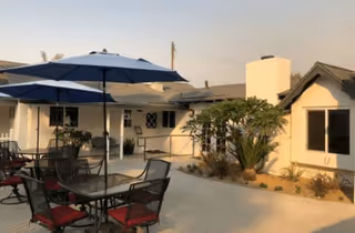 Outdoor patio area with tables and chairs featuring red cushions, shaded by large blue umbrellas. The patio is adjacent to a light-colored building with windows and a small garden with plants and shrubs. The sky is clear with a warm light indicating either morning or evening.