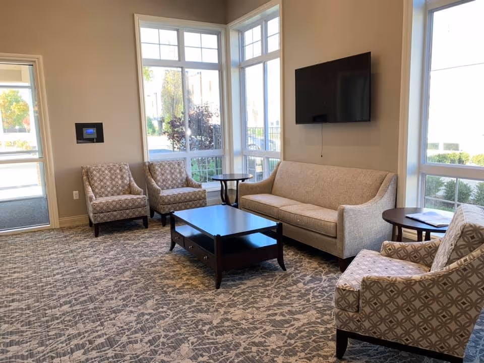 A bright and cozy sitting area in a senior living facility with large windows letting in natural light. The room features a beige sofa, three patterned armchairs, a dark wooden coffee table, and two small round side tables. A flat-screen TV is mounted on the wall above the sofa.