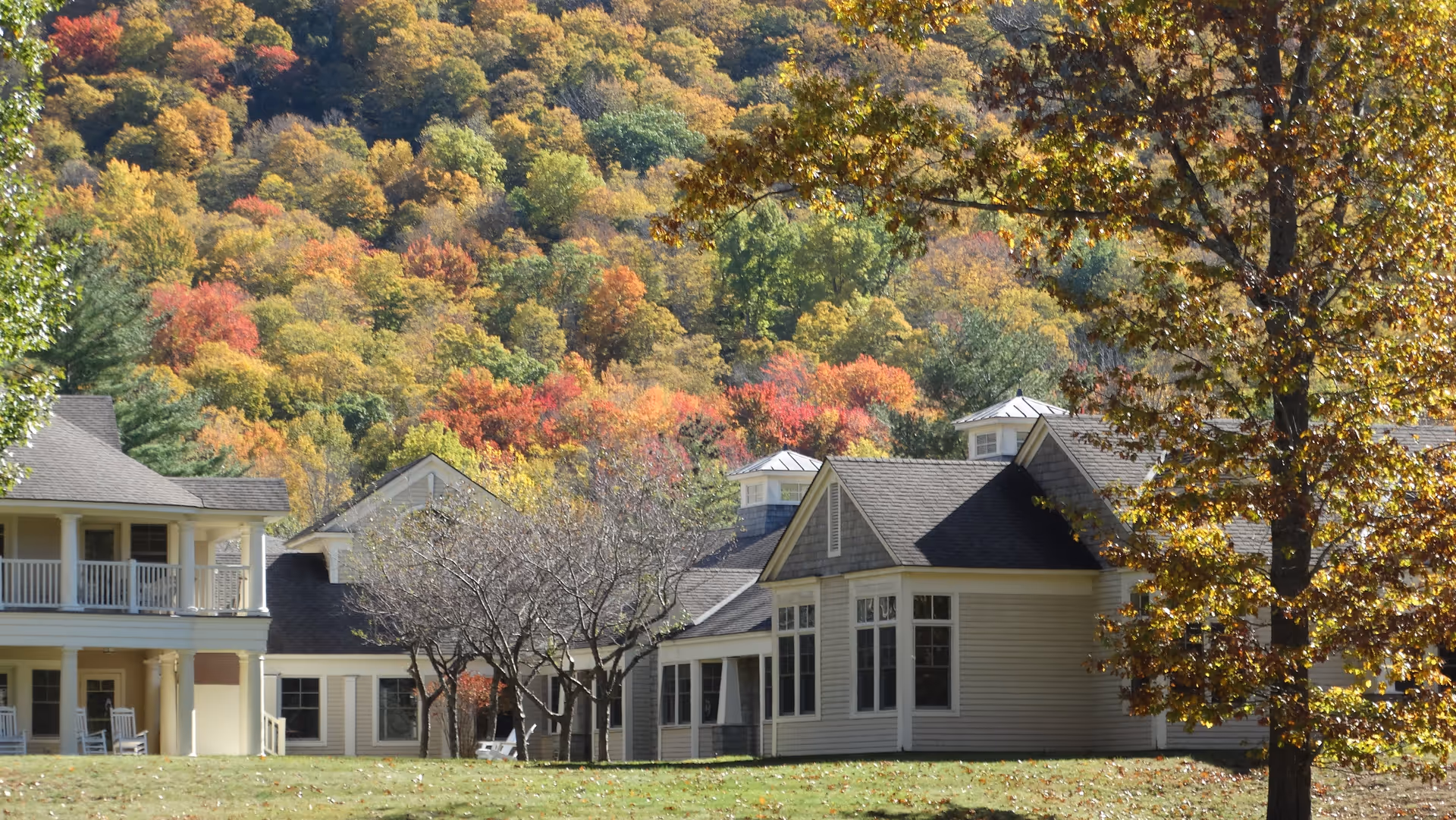 Cream-colored senior living building with porches and a lawn in front of a hillside covered in colorful autumn trees.