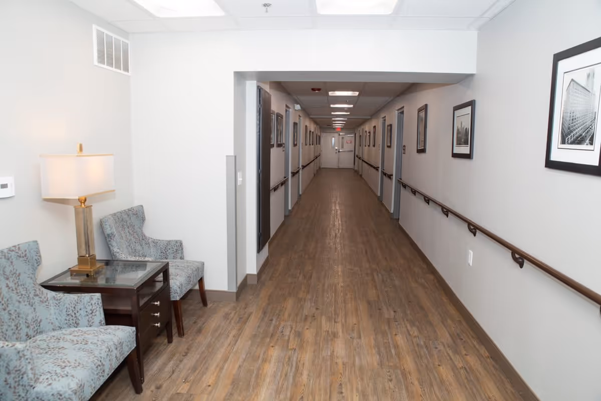 A long, well-lit hallway with wooden flooring and light gray walls. The hallway has handrails on both sides and framed black-and-white photographs hanging on the walls. On the left side near the foreground, there are two patterned armchairs and a small wooden table with a lamp on it.
