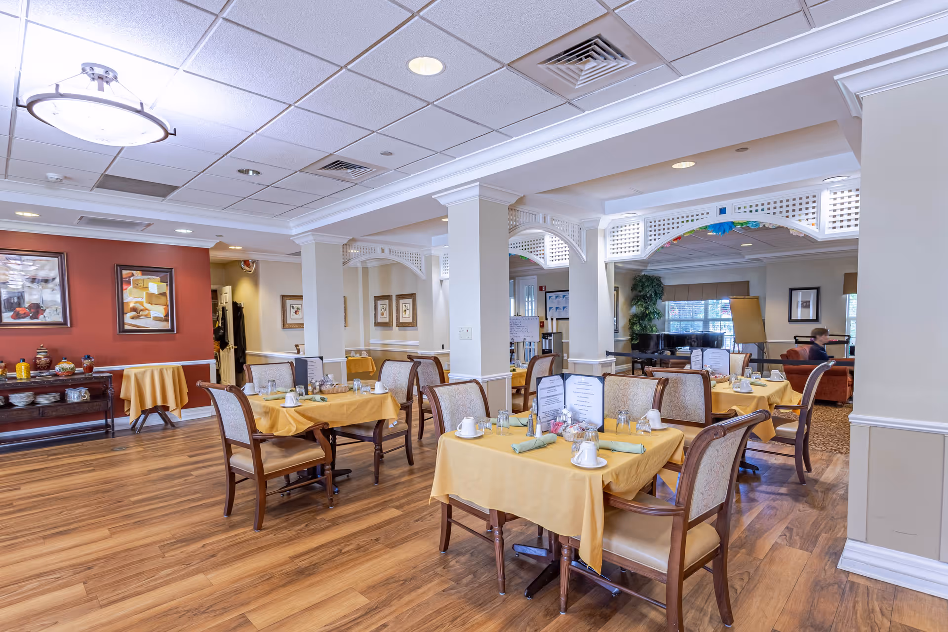 A bright and spacious dining room in Bear Creek Assisted & Senior Living with several tables covered in yellow tablecloths, each set with cups, napkins, and menus. The room features wooden flooring, white columns with decorative latticework, framed artwork on the walls, and a cozy seating area with a piano and large windows in the background.