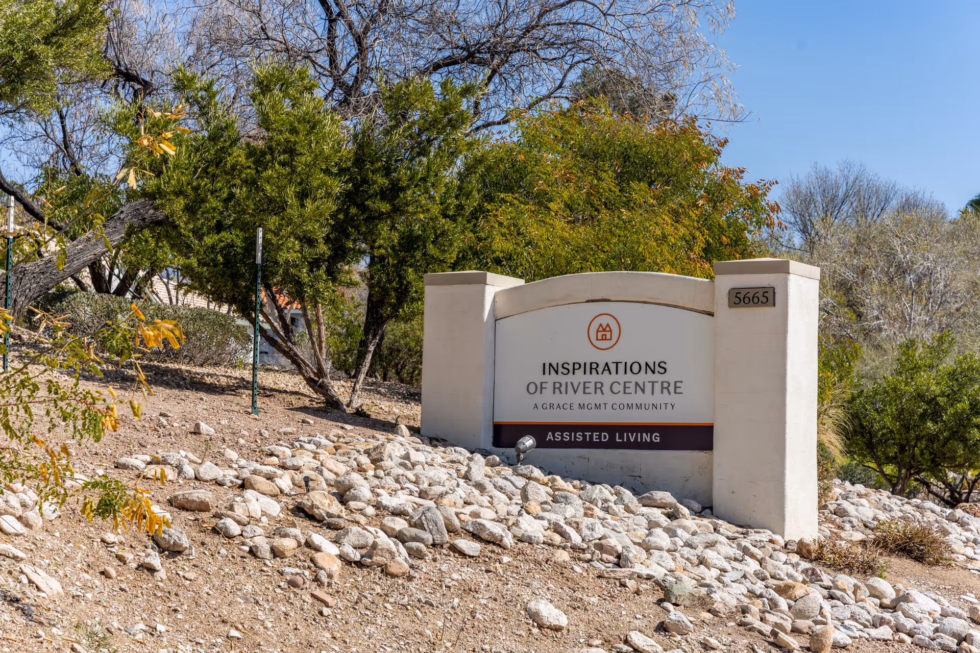 Outdoor view of a sign for Inspirations of River Centre, an assisted living community managed by Grace Management, surrounded by rocks and trees under a clear blue sky.