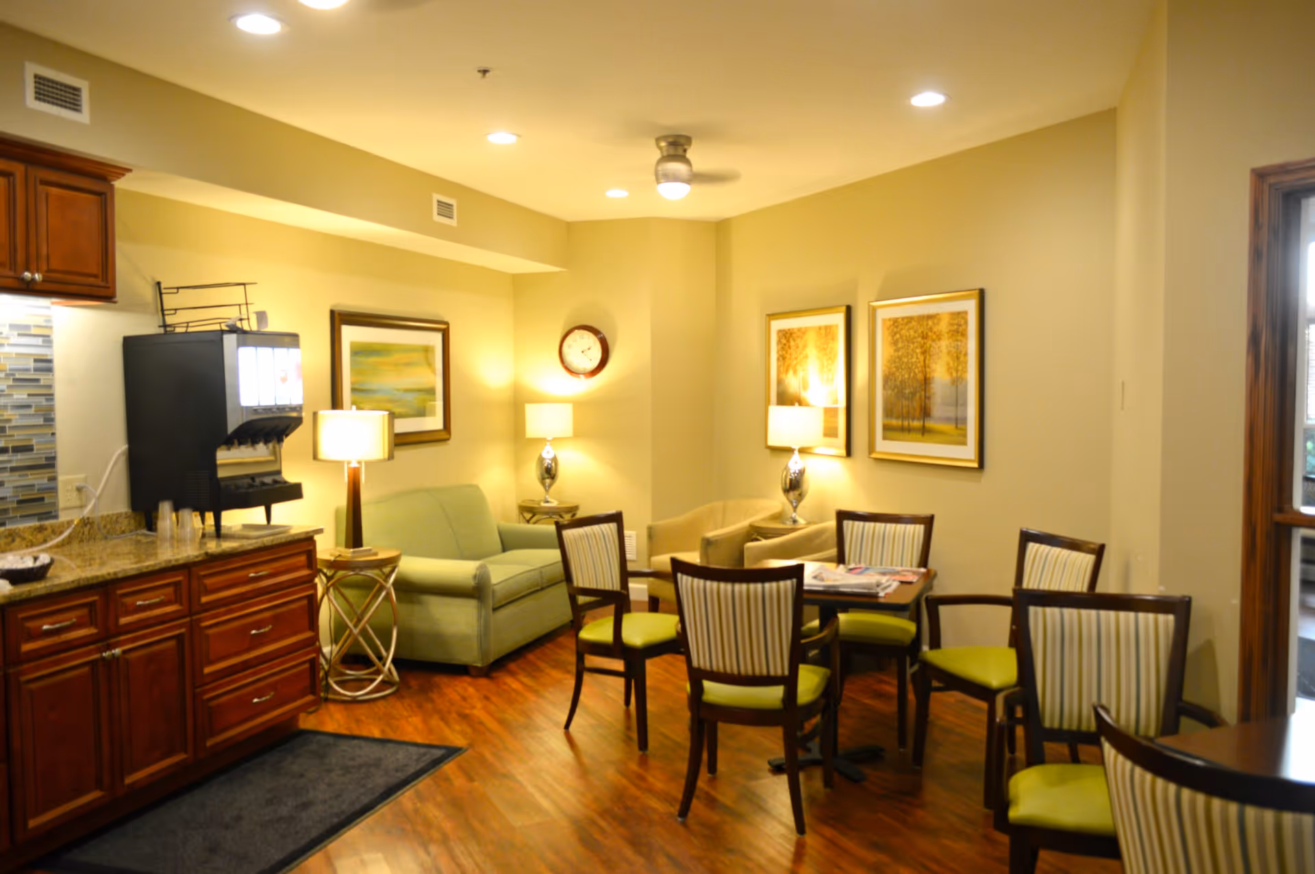 A cozy common area with wooden flooring, a green loveseat, two beige armchairs, a round table with four chairs featuring green cushions, and two framed pictures on the wall. There are two table lamps and a wall clock. To the left, there is a countertop with a beverage dispenser and wooden cabinets underneath.