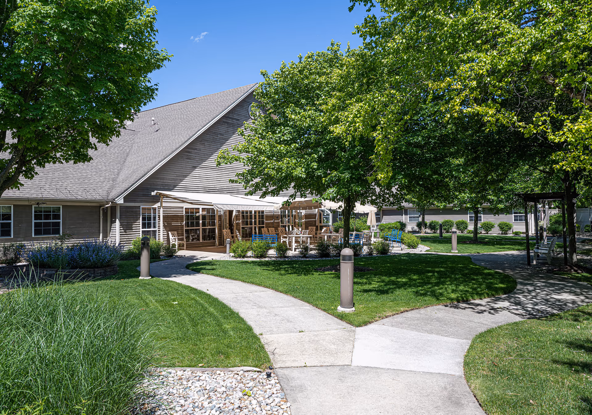 Outdoor view of a senior living facility with a paved walkway splitting into two paths surrounded by green grass, trees, and shrubs. There is a building with a covered patio area furnished with tables and chairs under a clear blue sky.