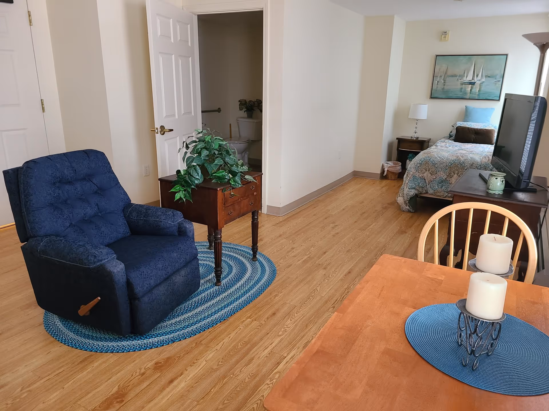 Studio-style senior living room with a navy recliner on a round blue rug, a small table with a plant, a dining table with candles, a TV stand, and a bed in the background.
