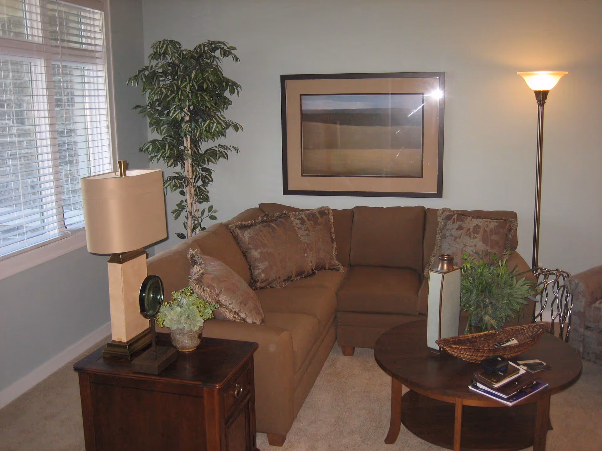 A cozy living room with a brown sectional sofa adorned with patterned cushions. A wooden side table with a beige lamp and a small plant sits next to the sofa. A tall potted plant is placed in the corner near a window with white blinds. A framed landscape picture hangs on the wall above the sofa. A round wooden coffee table in front of the sofa holds a decorative vase, a wicker basket, and some books. A floor lamp with a white shade is lit in the corner.