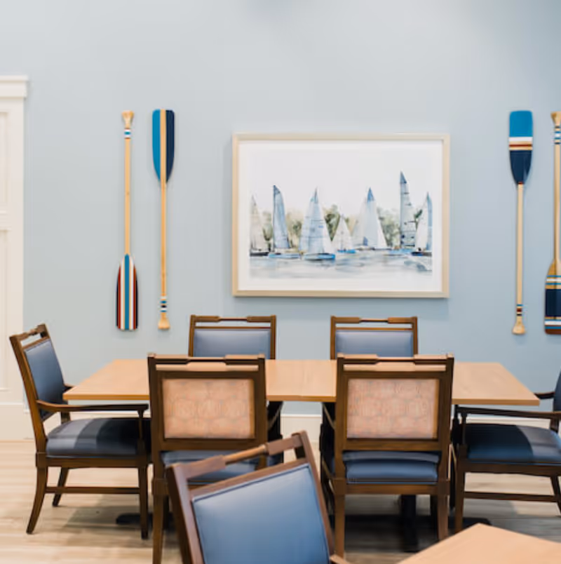 A dining room with a wooden table surrounded by six chairs with blue cushions and patterned backs. The wall behind the table is painted light blue and decorated with a framed painting of sailboats on the water and four decorative wooden oars mounted vertically on either side of the painting.