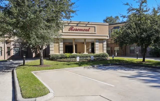 Front exterior view of the Rosemont Assisted Living And Memory Care facility with a driveway, parking spaces, green lawns, and trees under a clear blue sky.