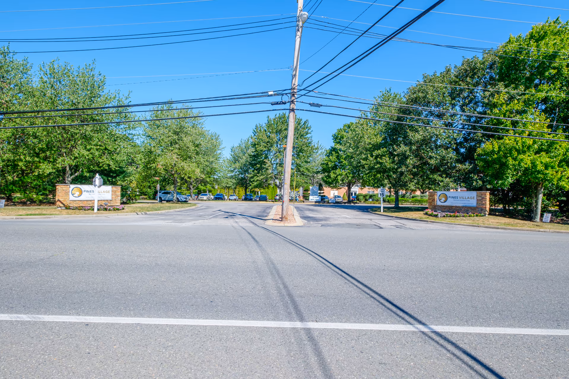 Entrance to Pines Village senior living facility with two stone signs on either side of the driveway, surrounded by green trees and a clear blue sky above. Several cars are parked inside the facility grounds.