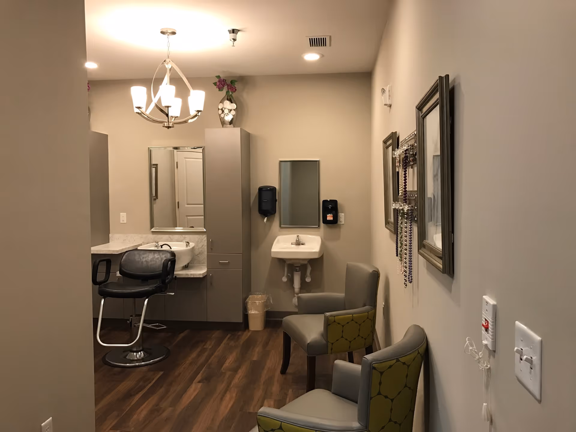 Interior view of a small salon or grooming area in a senior living facility. The room features a salon chair in front of a counter with a sink and a large mirror. There is a tall cabinet with a vase of flowers on top. On the right side, there is a wall-mounted sink with a mirror above it, a soap dispenser, and two upholstered chairs with patterned backs. The walls are decorated with framed pictures and a jewelry holder with hanging necklaces. The floor is wood, and the ceiling has recessed lighting and a chandelier.
