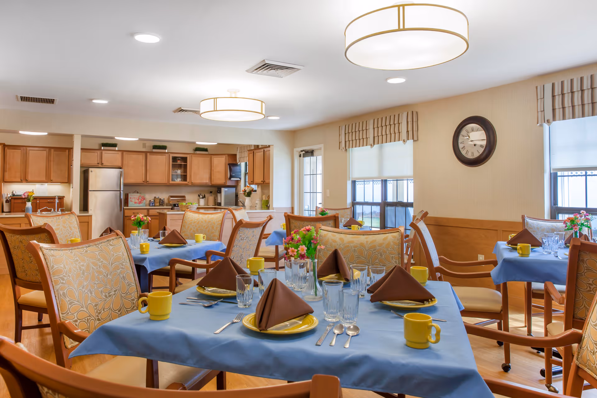A bright dining room in a senior living facility with several tables covered in blue tablecloths, set with yellow mugs, brown folded napkins, plates, glasses, and silverware. The room features wooden chairs with patterned cushions, large windows with striped valances, a wall clock, and a kitchen area with wooden cabinets and stainless steel appliances in the background.