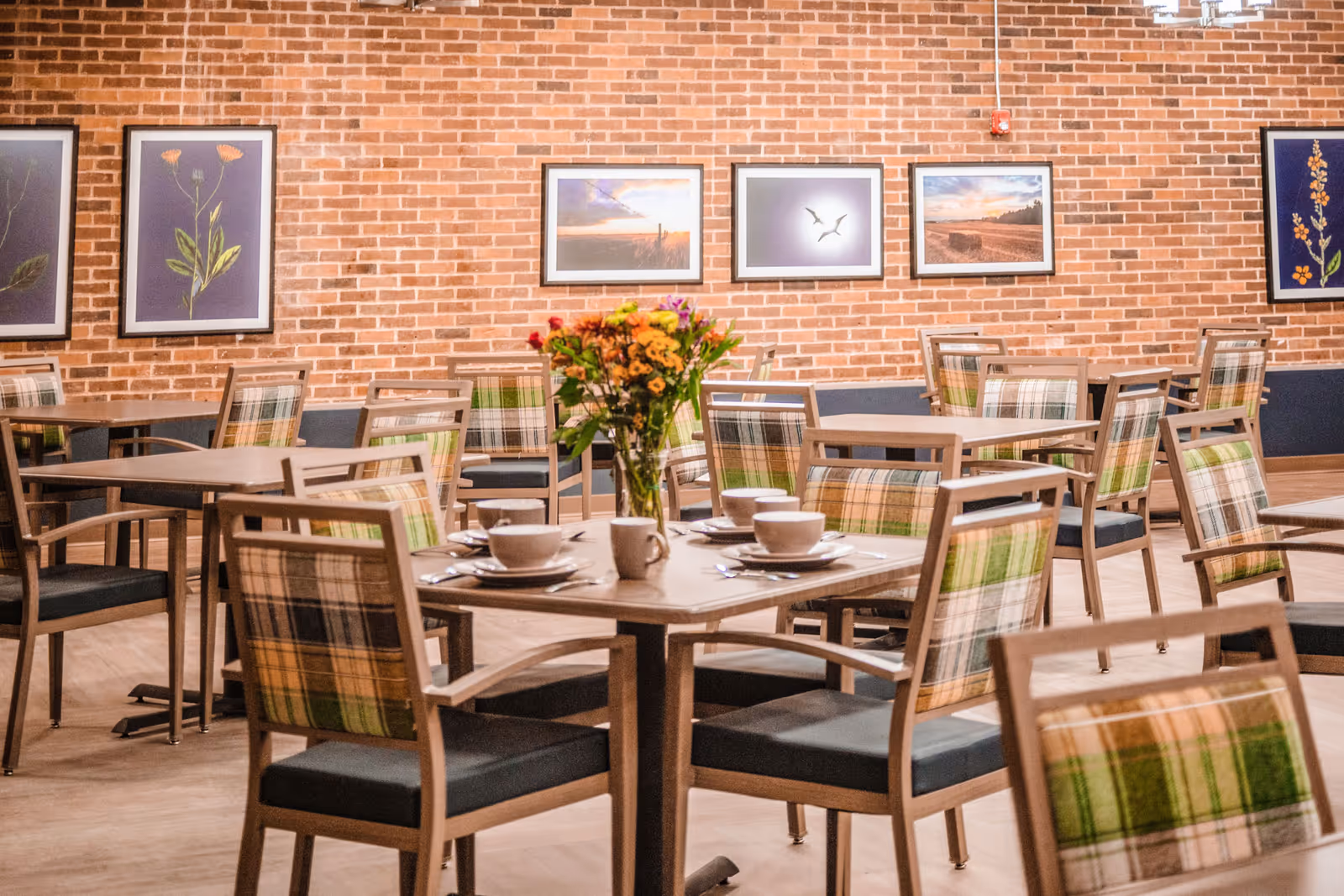 A dining room with multiple wooden tables and chairs featuring green and beige plaid upholstery. One table in the foreground is set with white cups, saucers, and a vase of colorful flowers. The background shows a brick wall adorned with framed pictures of flowers and nature scenes.