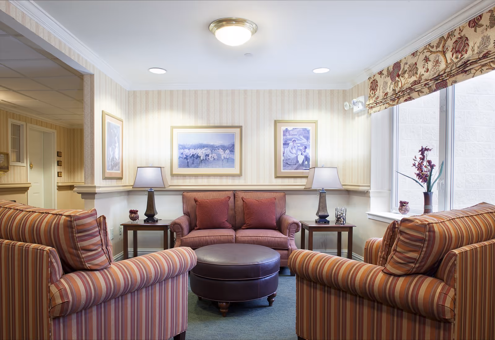 A cozy living room area with a striped armchair on each side, a small sofa with two red cushions in the center, and a round brown ottoman in front. Two side tables with lamps flank the sofa. The walls have light striped wallpaper and two framed pictures. A large window with floral curtains lets in natural light, and there are decorative plants on the window sill.
