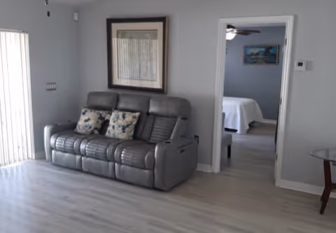 A living room area with a gray leather sofa featuring two floral cushions. There is a framed picture hanging on the wall above the sofa. To the right, an open doorway leads to a bedroom with a bed covered in white bedding and a painting on the wall. The floor is light-colored wood, and vertical blinds cover a window on the left side.