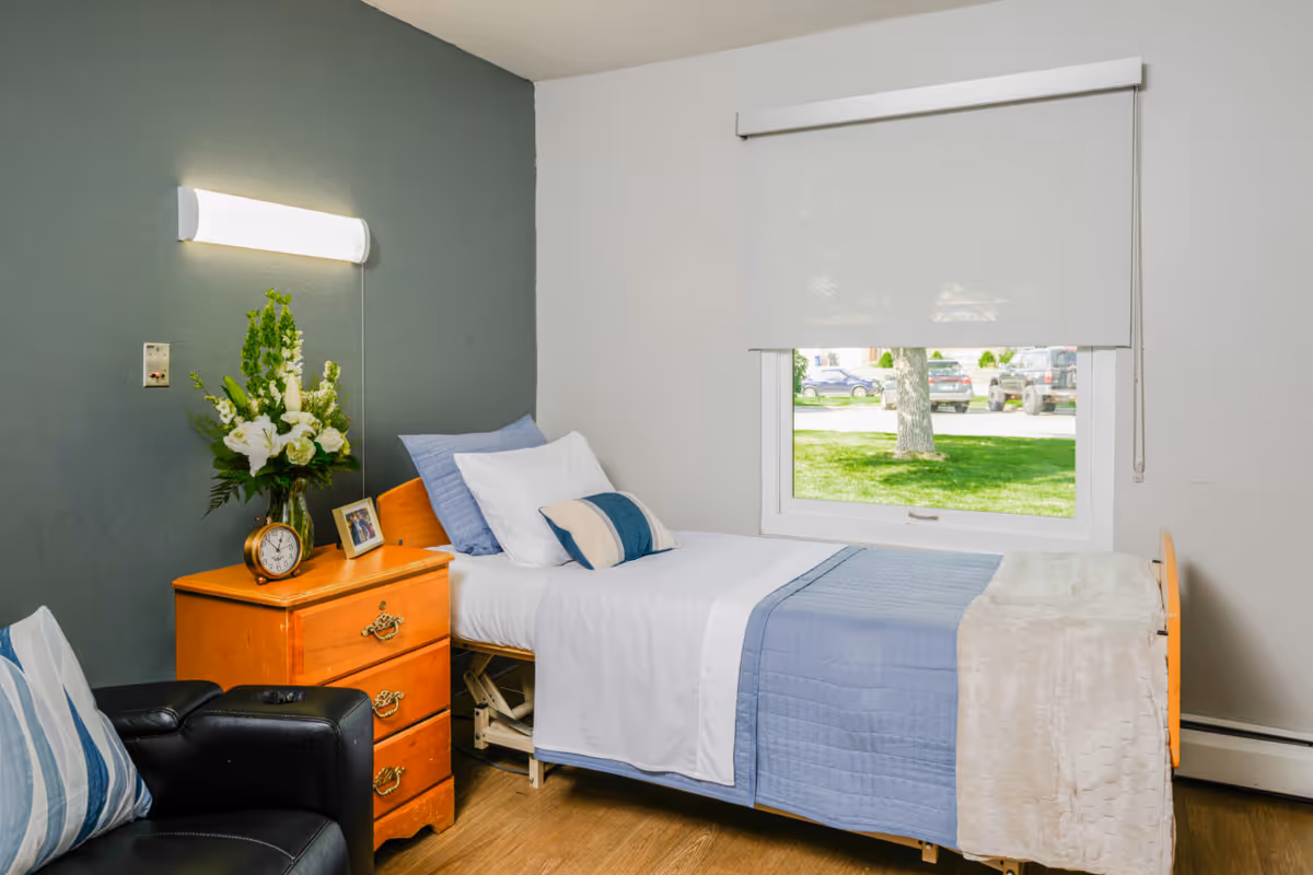 A neatly made bedroom with a single bed, wooden dresser, a vase of flowers, and a window with a shade.