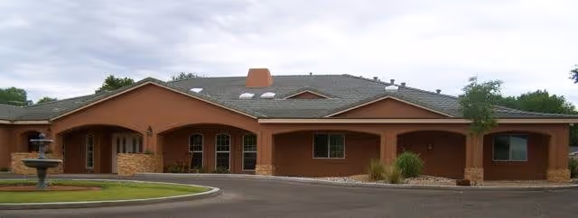 Single-story building with a brown exterior and a gray roof, featuring multiple windows and a covered entrance. There is a circular driveway in front with a fountain on the left side and some landscaping with grass and bushes.