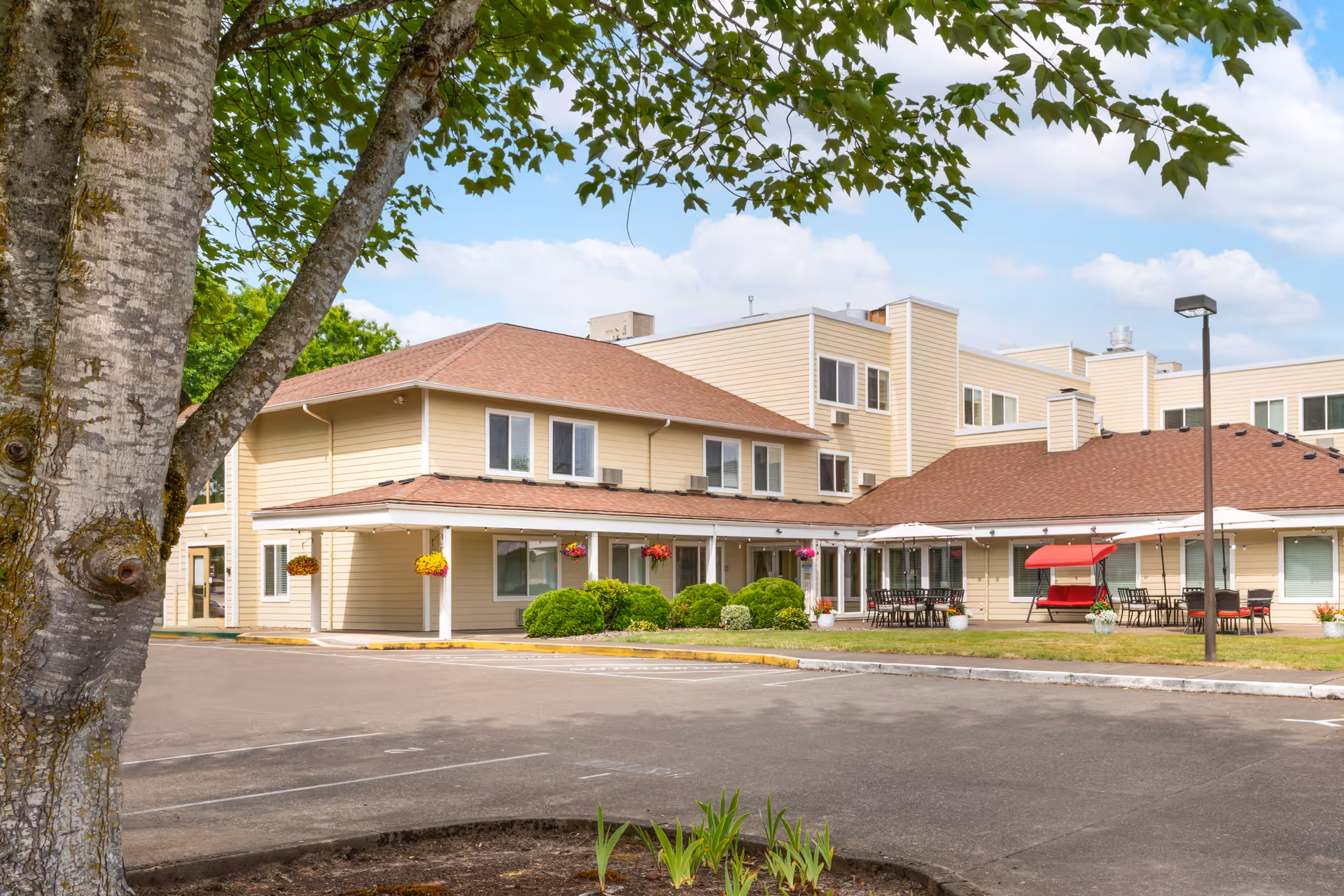 Exterior view of Brookdale Heritage Plaza, a beige two-story building with a red roof, surrounded by greenery and outdoor seating areas with tables, chairs, and umbrellas. A large tree is visible in the foreground with a clear blue sky and some clouds in the background.