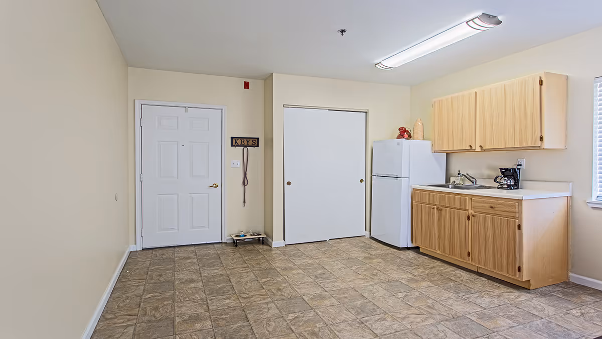 Interior view of a small kitchen area in a senior living facility with a white entrance door, a white refrigerator, wooden cabinets, a sink, a coffee maker, and a tiled floor. There is a key holder on the wall next to the door and a window on the right side letting in natural light.