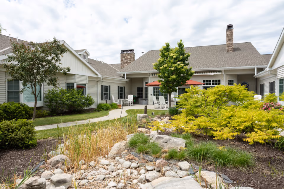 Outdoor garden area at CountryHouse Residence featuring a landscaped courtyard with various green shrubs, small trees, rocks, and a stone pathway leading to a building with beige siding and multiple windows. There are white chairs and red umbrellas on a patio area near the building under a partly cloudy sky.