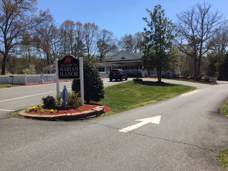 Driveway leading to the white Marian Manor entrance with a welcome sign, landscaped island, and a parked car.