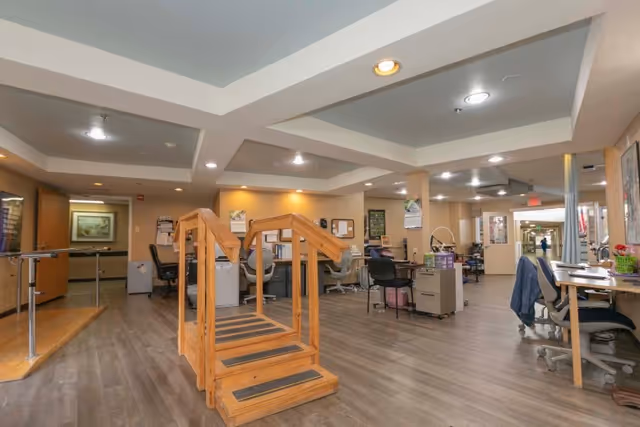 Open rehabilitation/activity room with wooden therapy stairs, parallel bars, desks and chairs under recessed ceilings.