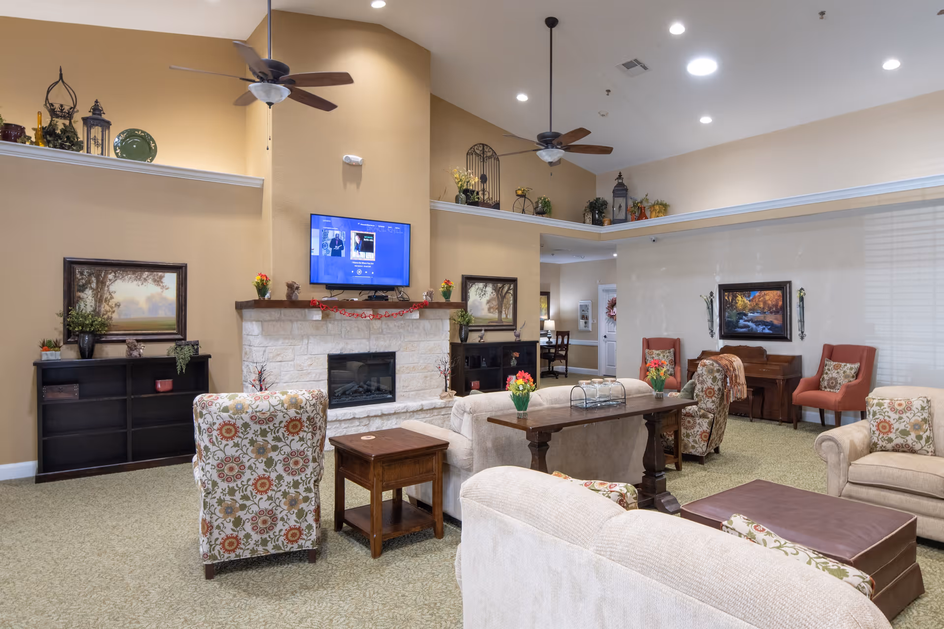 A spacious senior living facility common area with beige walls and carpet. The room features a stone fireplace with a mounted TV above it, two ceiling fans, and various seating options including floral patterned armchairs, beige sofas, and coral-colored chairs. There are decorative plants, paintings on the walls, and a piano in the corner. The room is well-lit with recessed lighting.