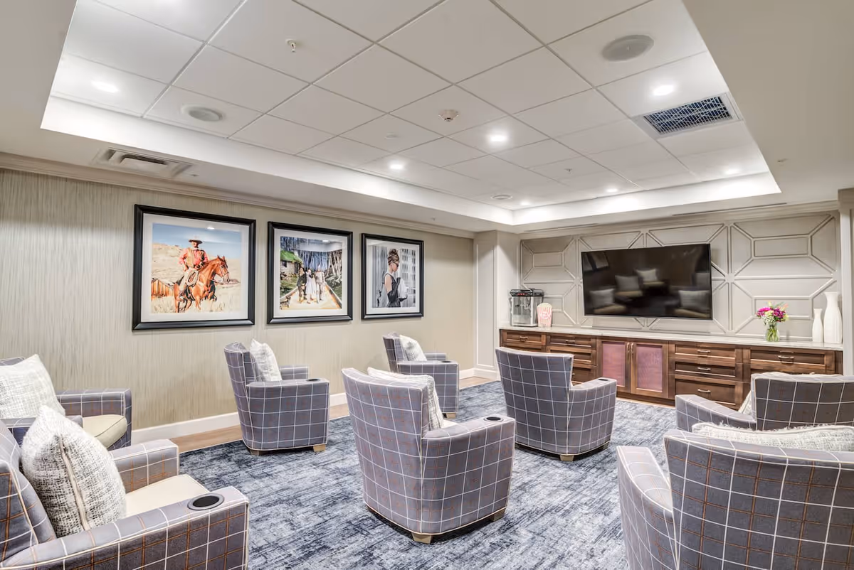 A cozy lounge area with six plaid armchairs arranged in a semi-circle facing a large flat-screen TV mounted on a wall with decorative paneling. Below the TV is a wooden cabinet with multiple drawers and a popcorn machine on top. The room has a blue patterned carpet, three framed pictures on one wall, and a vase with flowers on the cabinet.