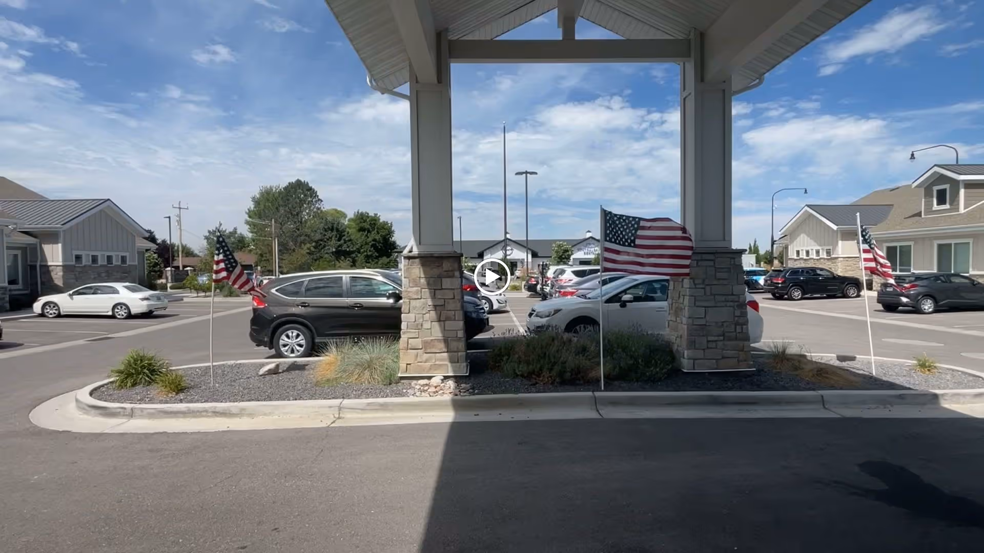 View of the entrance driveway and parking area of a senior living facility with stone pillars supporting a covered drop-off area. Several American flags are displayed on flagpoles along the landscaped median. Multiple cars are parked in the background near residential-style buildings under a partly cloudy sky.