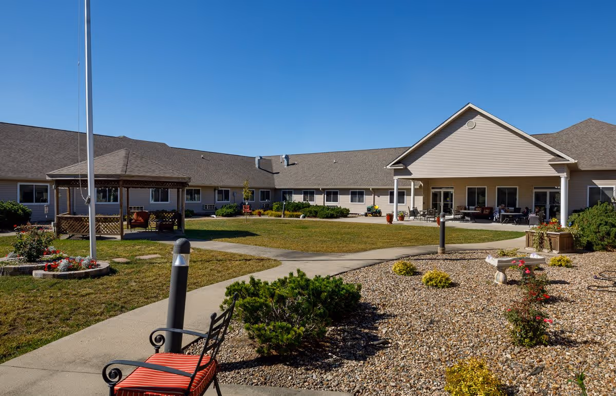 Outdoor courtyard area of Homestead Assisted Living & Memory Care of Oskaloosa featuring a paved walkway, a gazebo with seating, landscaped garden beds with flowers and shrubs, and a building with a covered patio and outdoor furniture under a clear blue sky.