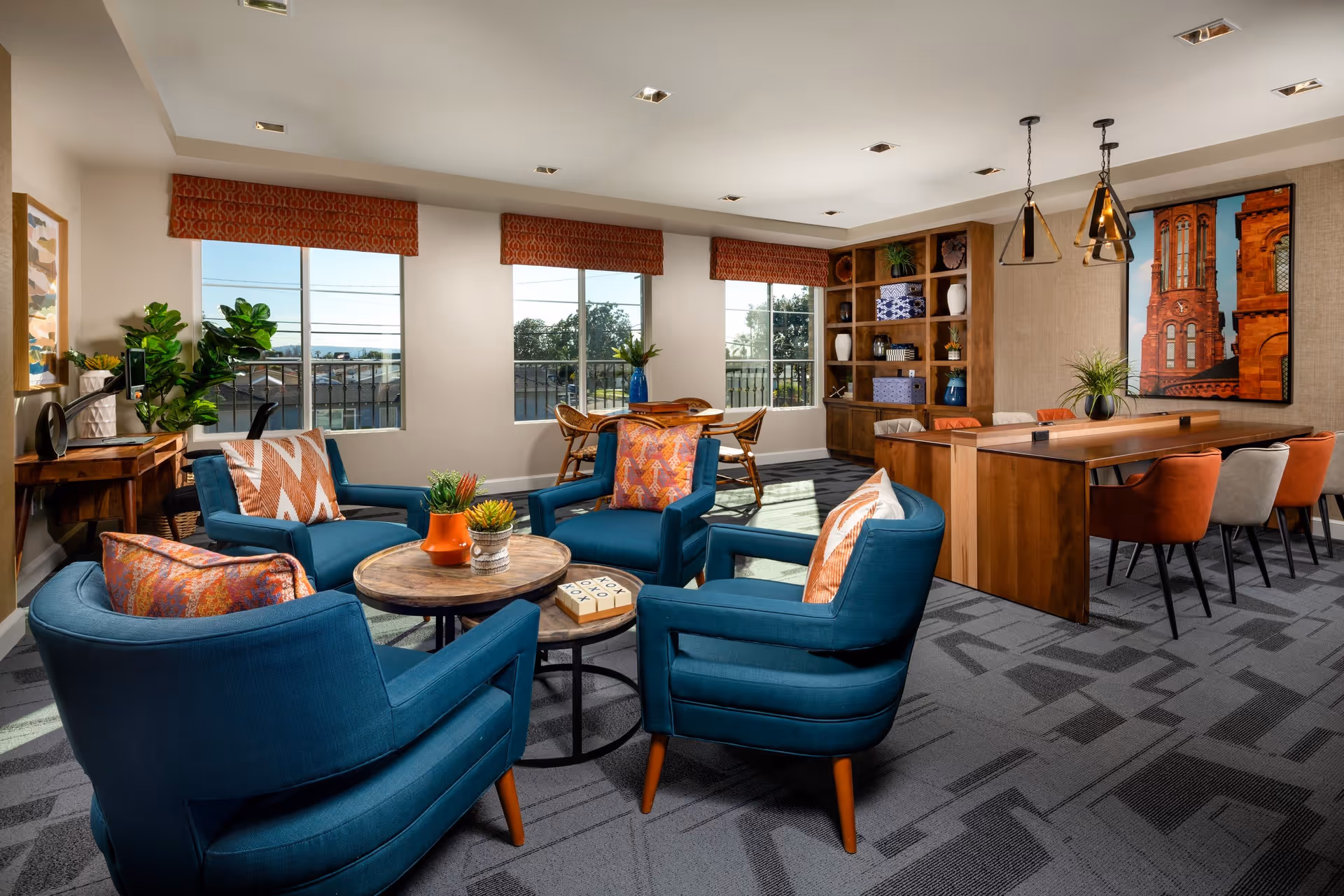 A bright and modern common area in a senior living facility featuring four blue armchairs with patterned orange and white pillows arranged around two round wooden coffee tables with small potted plants. Behind the seating area, there is a wooden table with six chairs in orange and beige colors, pendant lights hanging above, and a large framed picture of a red brick building on the wall. The room has three large windows with orange valances, a wooden shelving unit with decorative items, and a small desk with a plant and lamp near the left wall.