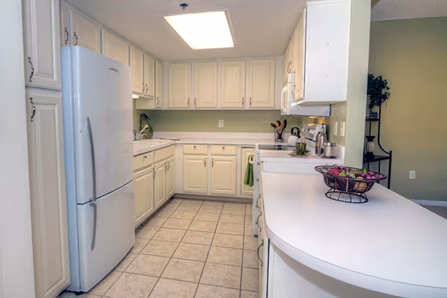 Bright white U-shaped kitchen with tiled floor, refrigerator, ample cabinets, and a curved breakfast bar with a fruit basket.