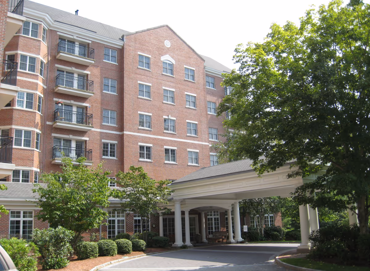 Exterior view of a multi-story brick senior living facility with balconies, large windows, and a covered entrance surrounded by trees and shrubs.
