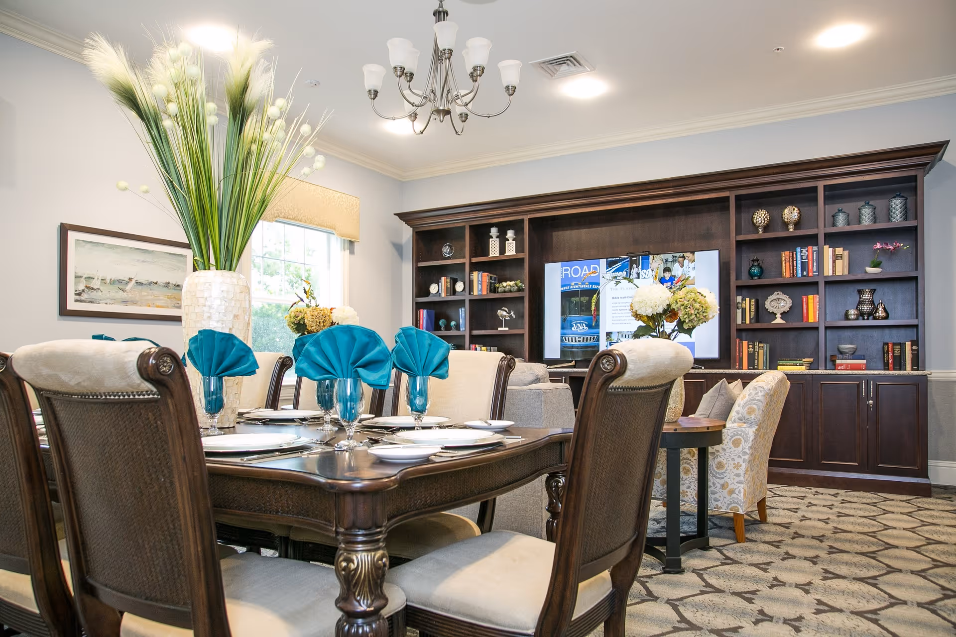 A well-decorated dining area with a wooden dining table set for six, featuring blue folded napkins in glasses. Behind the dining area is a living space with a large dark wood built-in bookshelf filled with books and decorative items, and a TV in the center. The room has a window with a beige valance, a chandelier, and a patterned carpet.