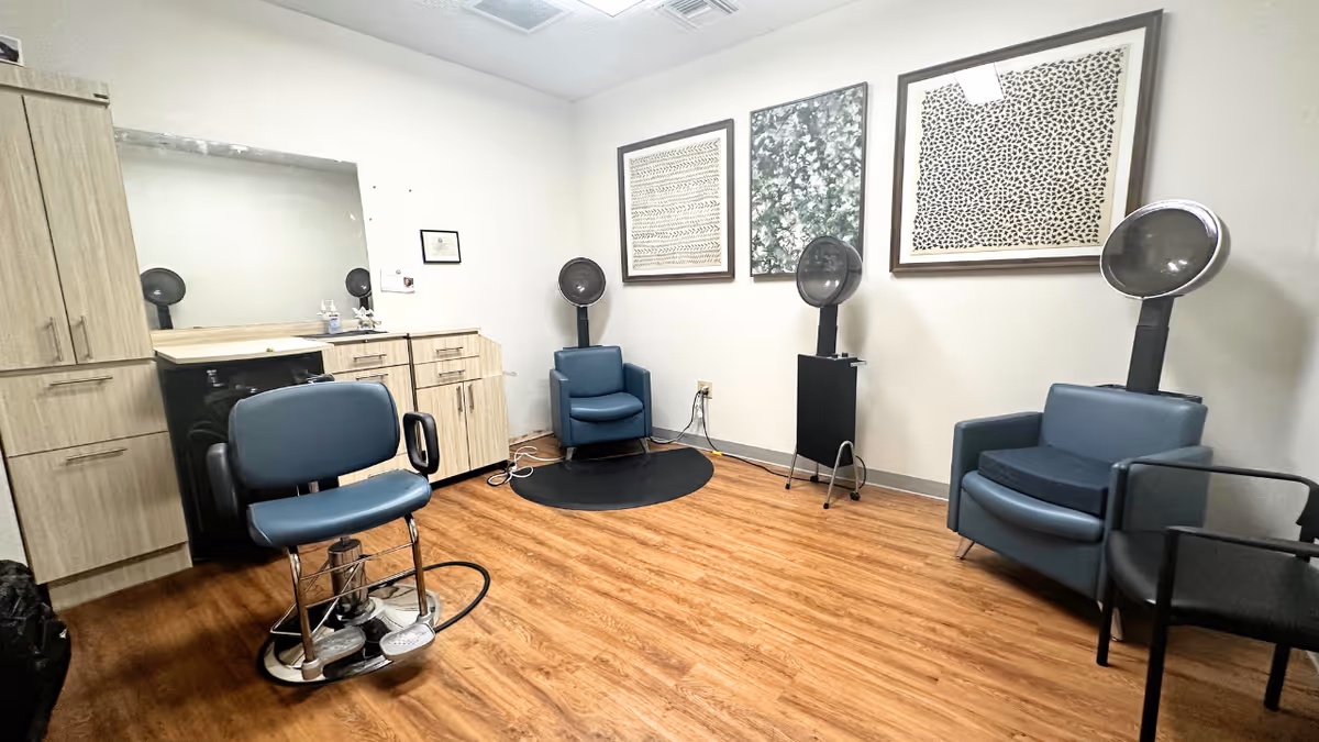 Small salon room with blue styling chairs, hooded hair dryers, cabinets, and framed artwork on the walls.