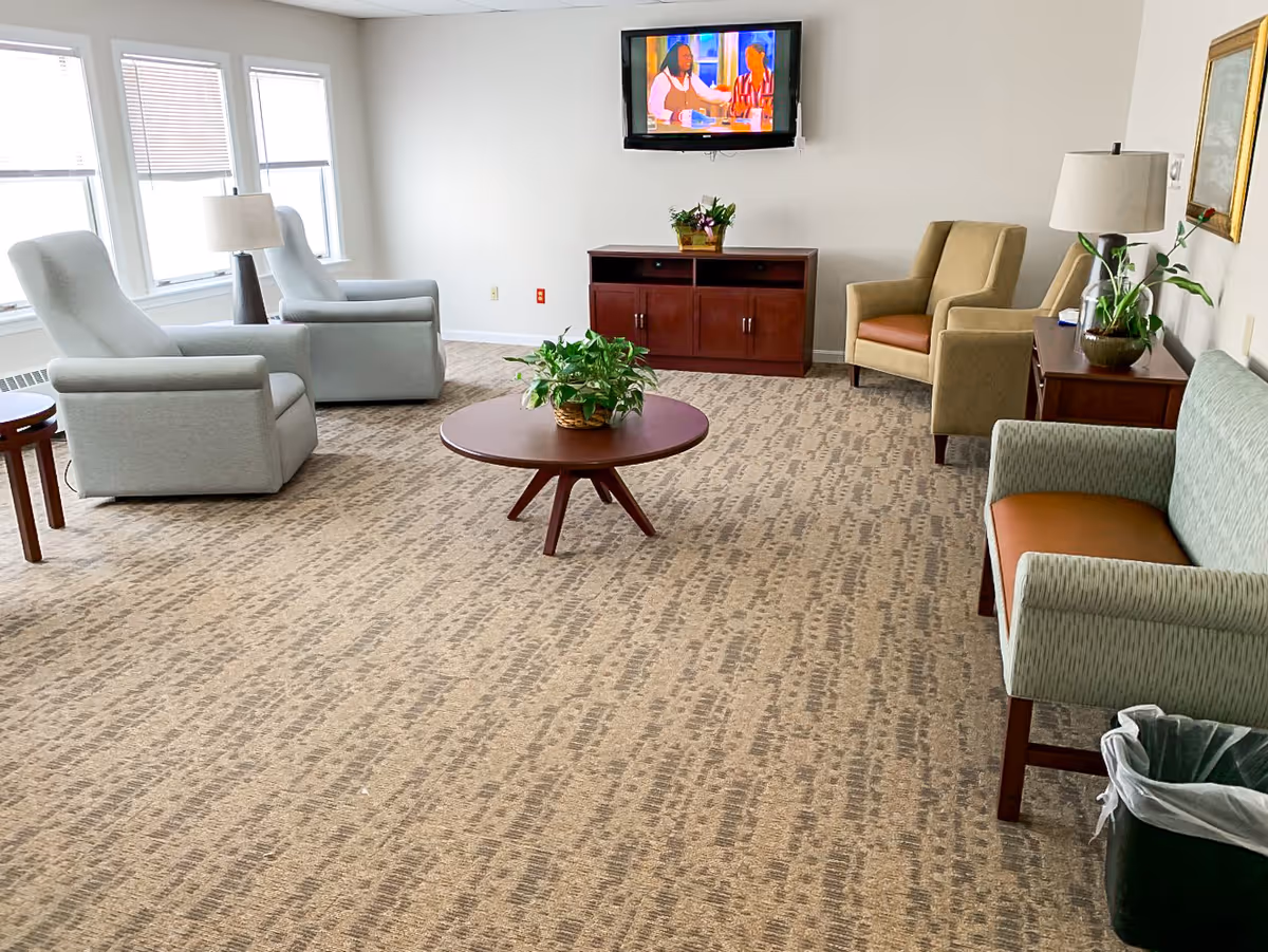 Bright communal lounge with armchairs, a sofa, a round coffee table with a plant, and a wall-mounted TV.