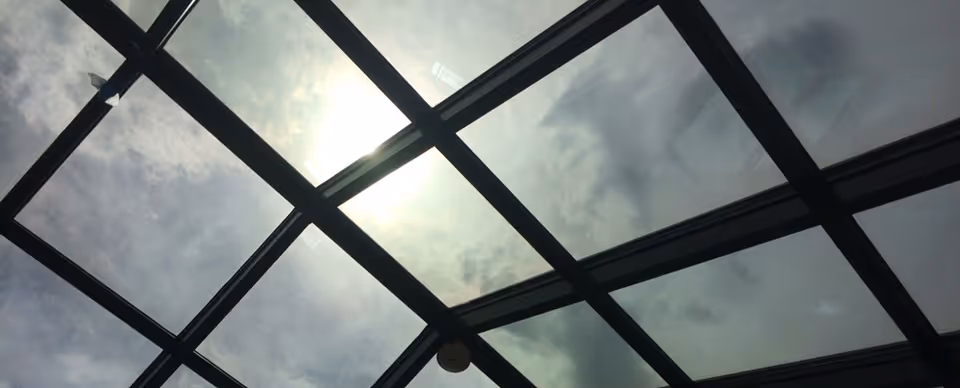 A large glass skylight with a dark metal grid framing a cloudy sky and the sun shining through.