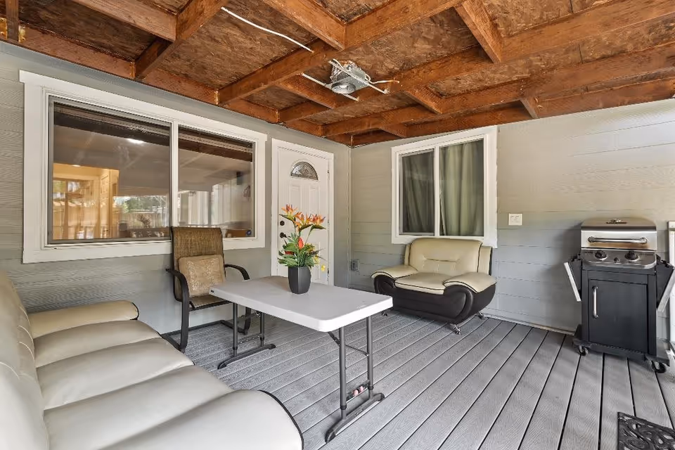 Covered outdoor patio area with a wooden ceiling, gray flooring, and gray walls. The space includes a white cushioned sofa, a cushioned armchair, a chair with a cushion, a white folding table with a potted plant on it, and a black grill on wheels.