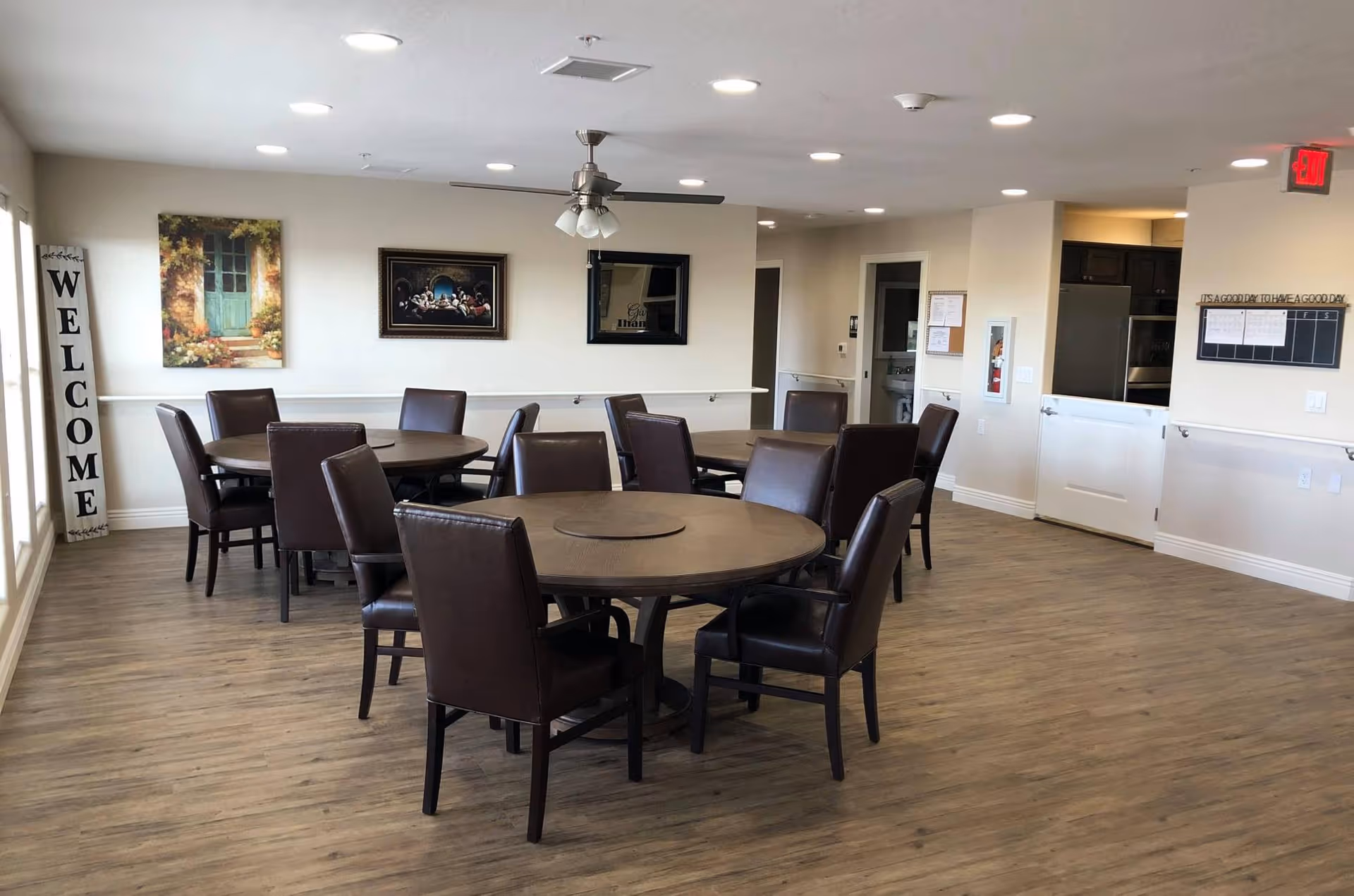 A spacious dining room with round tables and brown leather chairs arranged neatly. The room has wood flooring, a ceiling fan with lights, and recessed ceiling lights. On the left wall, there is a vertical 'WELCOME' sign and two framed pictures. The right side shows a partial view of a kitchen area and an exit sign above a door.