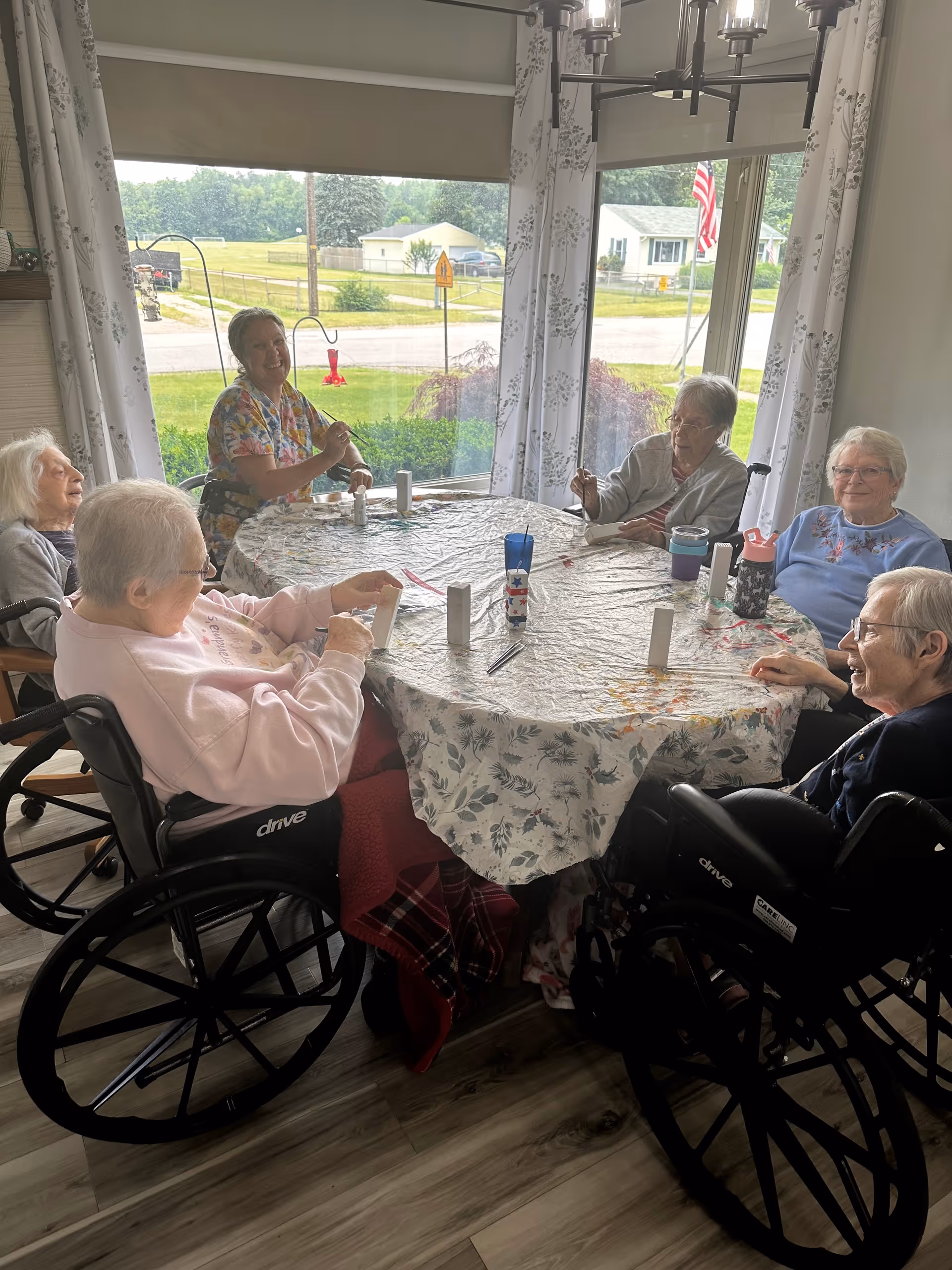 A group of elderly women sitting around a round table covered with a floral tablecloth in a bright room with large windows. Some women are in wheelchairs, and they appear to be engaged in a craft or activity involving small white objects. A caregiver or staff member is also seated at the table, smiling. Outside the windows, a suburban neighborhood with houses and greenery is visible.