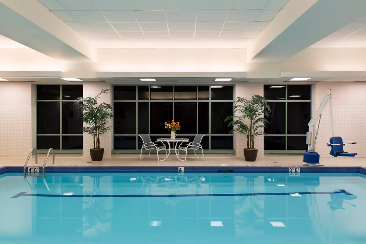 Indoor swimming pool with clear blue water, a metal pool ladder on the left, a pool lift chair on the right, two potted plants flanking a small round table with four chairs and a vase of flowers in the center, large windows in the background.