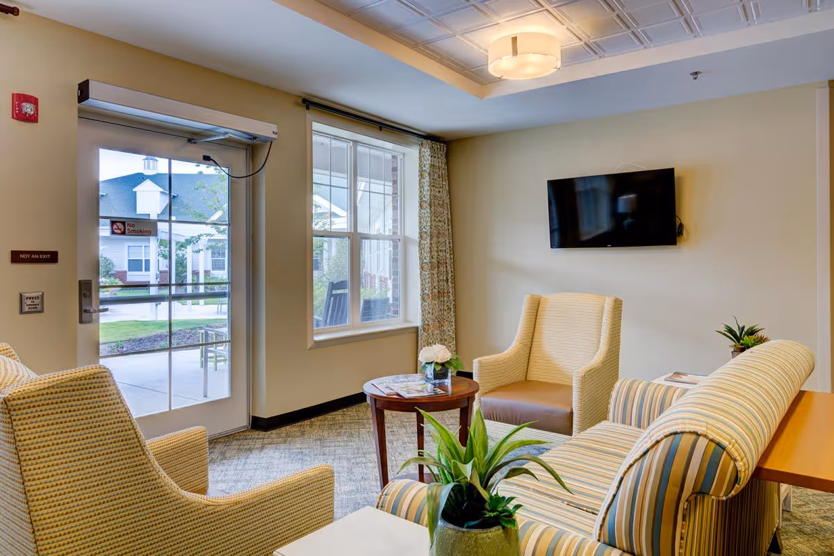 A cozy seating area in a senior living facility with two striped armchairs, a striped sofa, a small round wooden table with a flower vase and magazines, a potted plant on a side table, a wall-mounted flat screen TV, and a glass door leading outside with a 'No Smoking' sign visible.