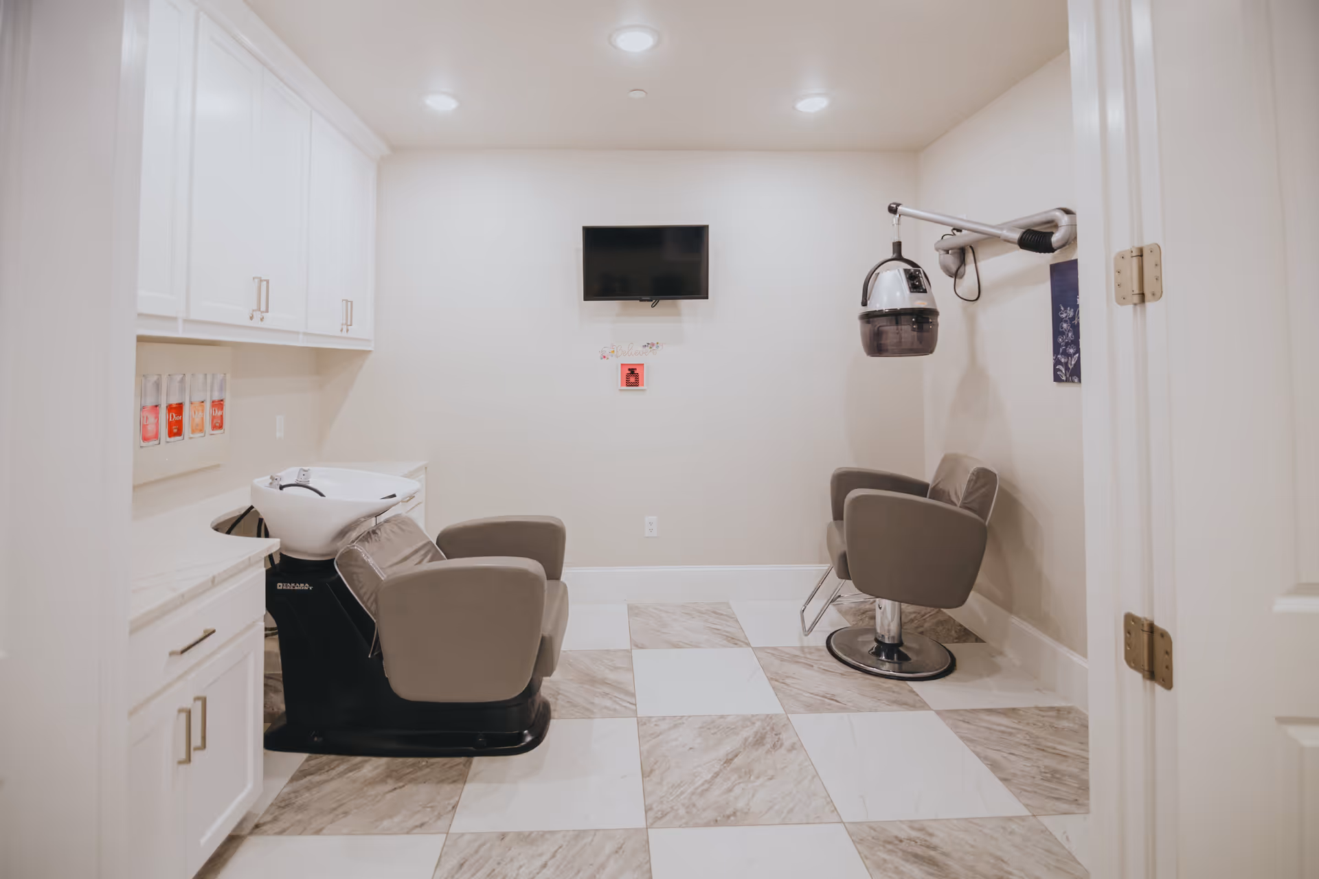 Interior view of a hair salon area in Avalon Memory Care featuring two gray salon chairs, a hair washing station with a white sink, a wall-mounted hair dryer, white cabinetry, and a flat-screen TV mounted on a beige wall.
