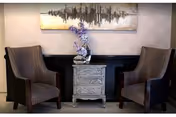 Two upholstered armchairs flank a small ornate side table topped with a vase of purple flowers beneath an abstract wall painting.