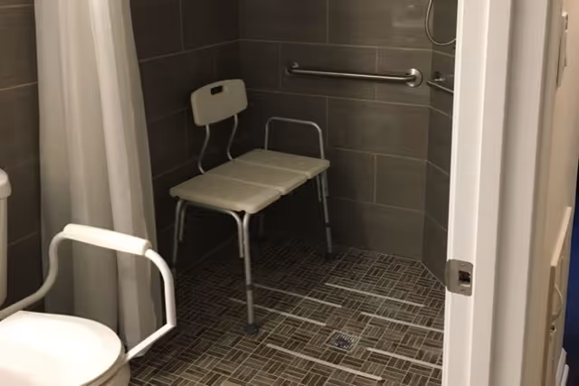Accessible bathroom with a white shower chair, grab bar, tiled walls and floor, and a toilet with arm support.