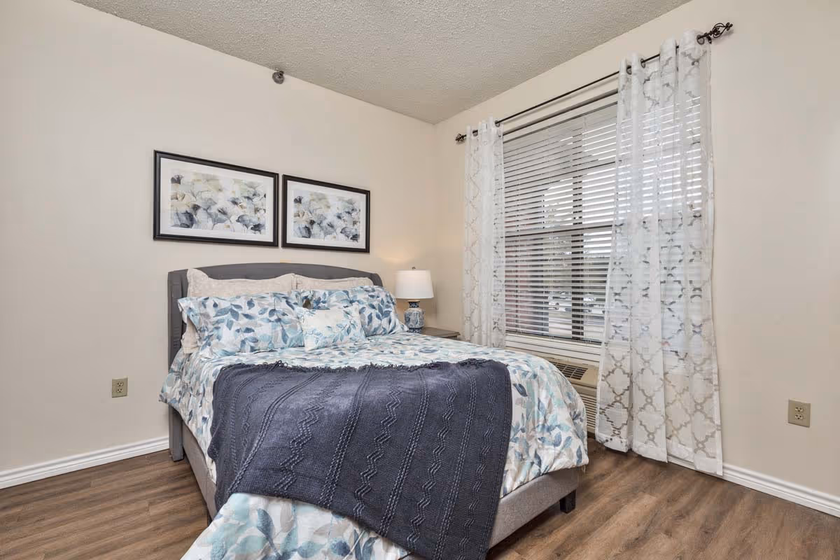 A neatly made bed with floral patterned bedding and a dark gray throw blanket in a bedroom. There are two framed floral artworks on the wall above the bed, a bedside table with a lamp, a window with white blinds and sheer patterned curtains, and wood flooring.