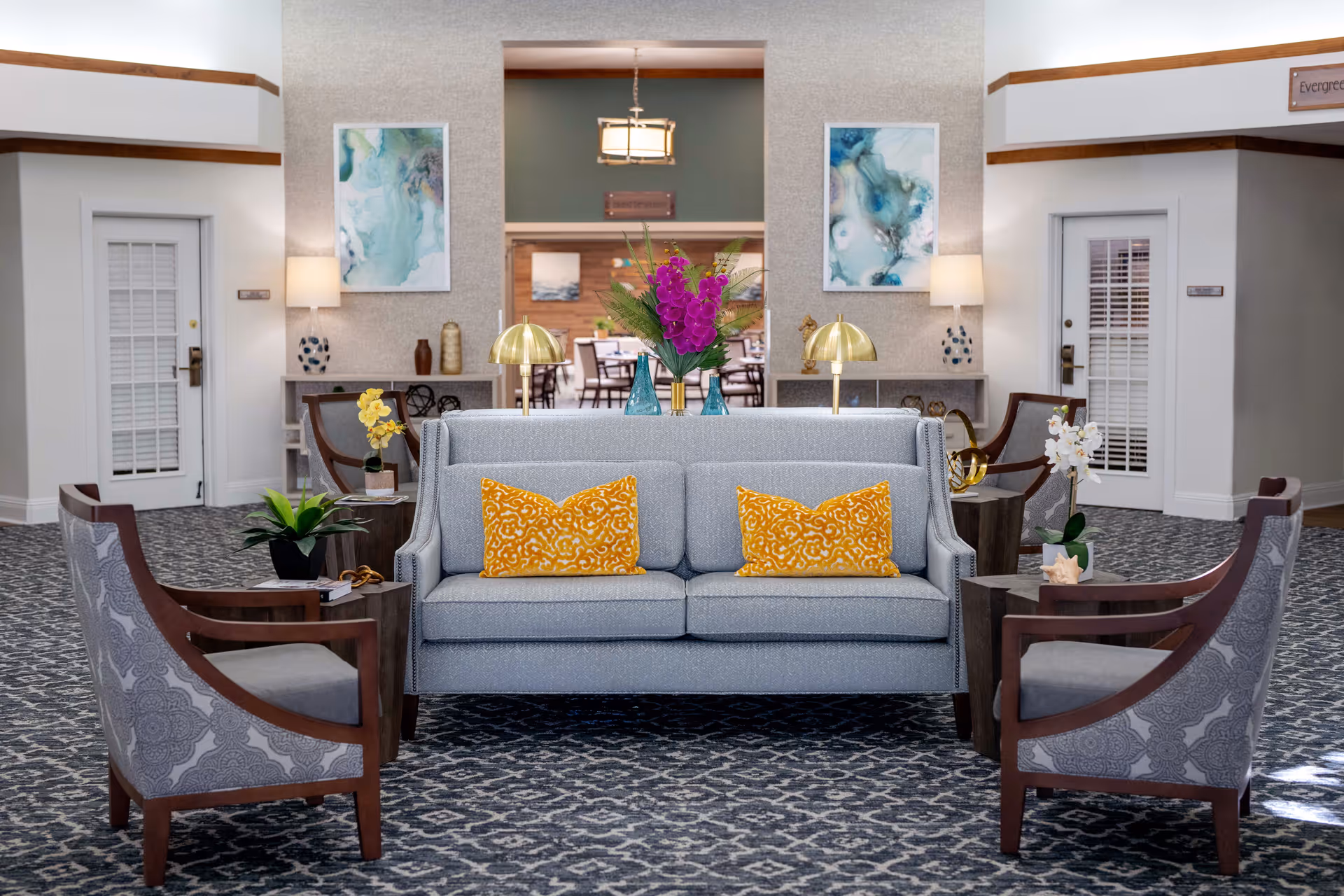A well-decorated senior living facility common area with a light gray sofa adorned with two yellow patterned pillows, surrounded by four wooden armchairs with gray patterned cushions. The room features a patterned carpet, side tables with plants and flowers, two table lamps with gold shades, and abstract artwork on the walls. In the background, there is a dining area with tables and chairs visible through a large open doorway.