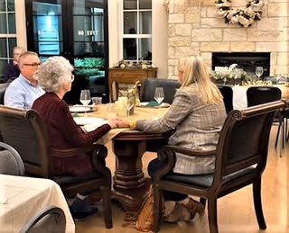 Three older adults seated around a dining table in a cozy room with a stone fireplace, wreath, and place settings.